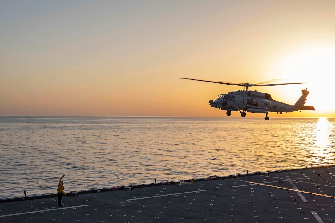 A sailor on a deck signals an airborne helicopter amid a golden sky.