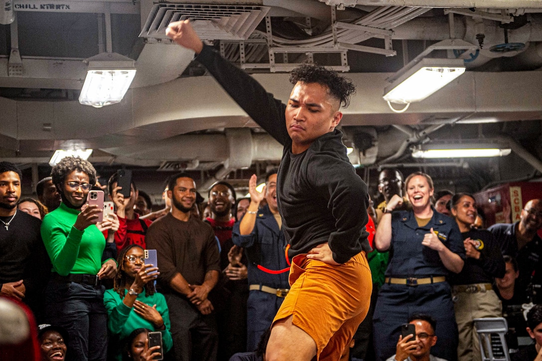 A sailor dances in an interior space on a ship as others cheer him on.