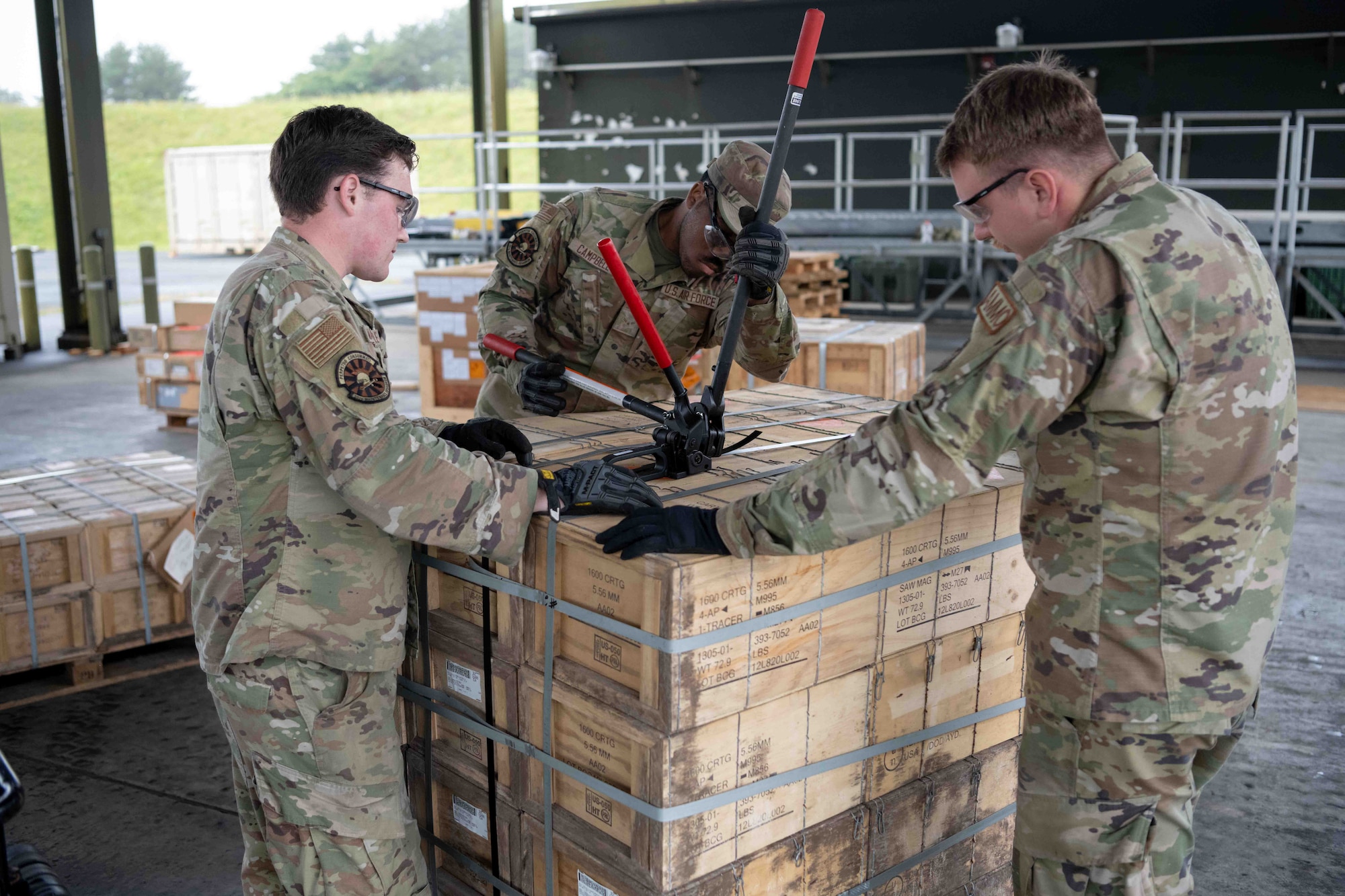 A military member tightens a metal band around a pallet to secure it and makes sure nothing falls off.