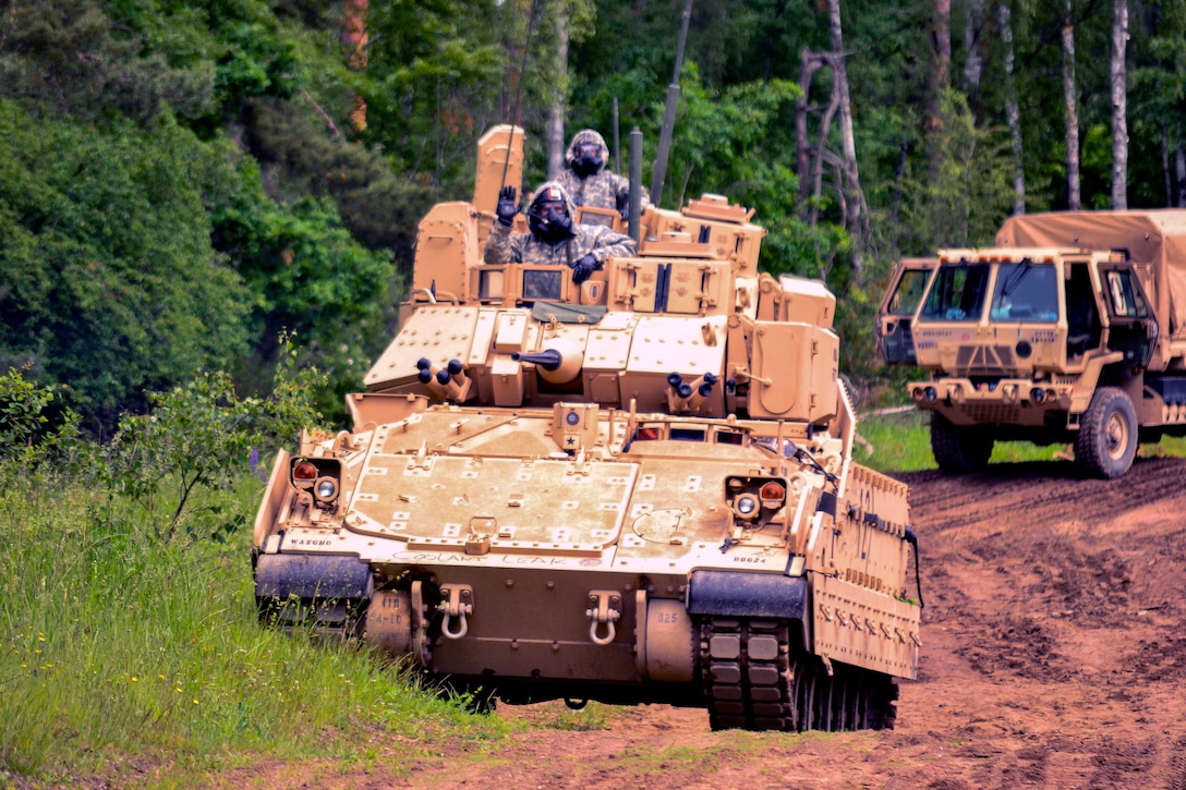 Several service members wearing chemical masks ride in tanks down a hill.