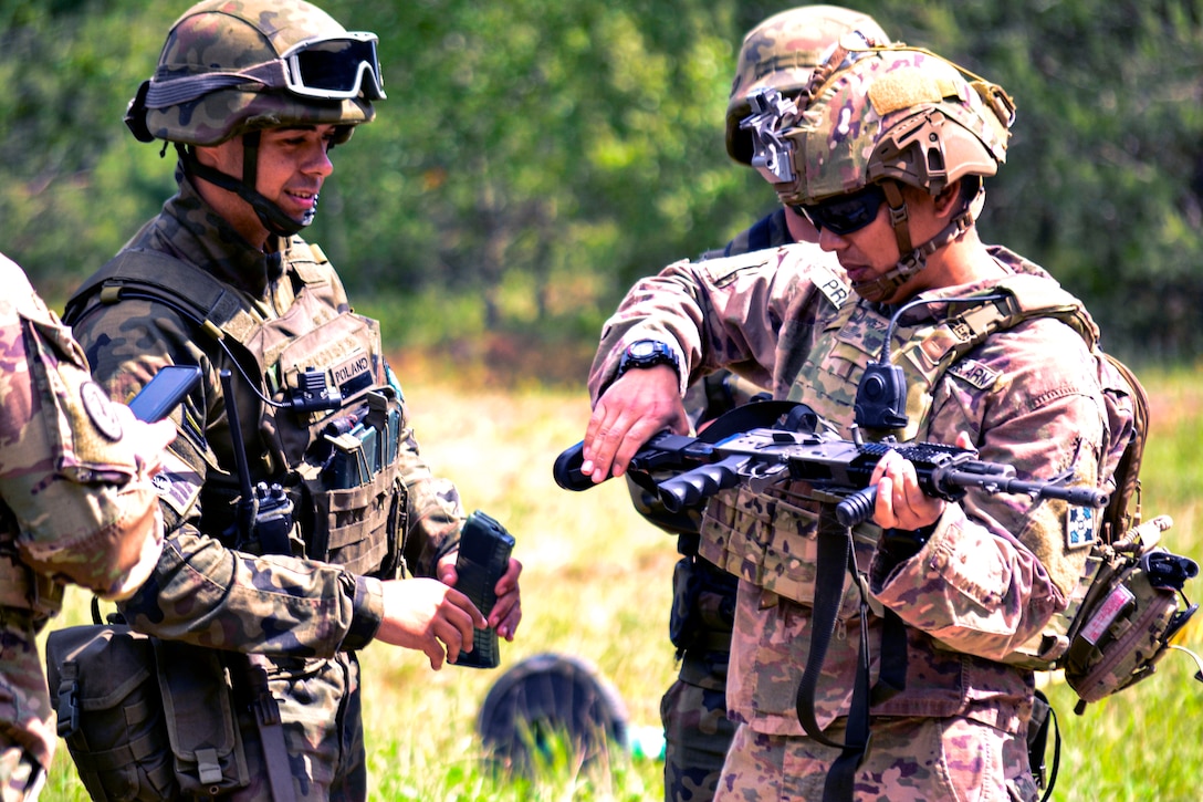 Four soldiers stand in a field watching one soldier manipulate a weapon.