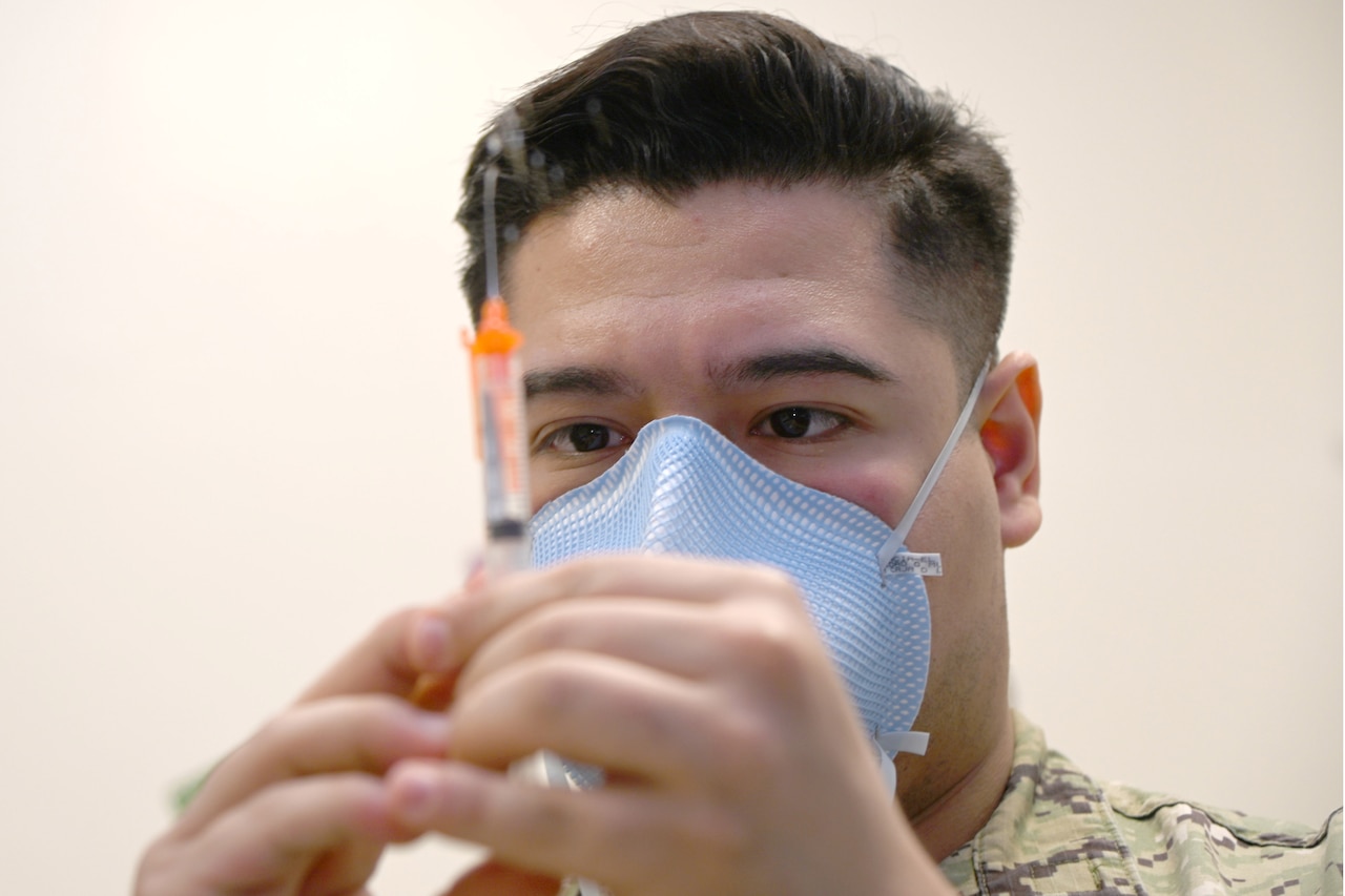 A sailor in a face mask looks at a syringe he's holding needle-side up.
