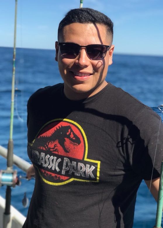 A sailor in civilian clothes and sunglasses smiles in front of a body of blue water.