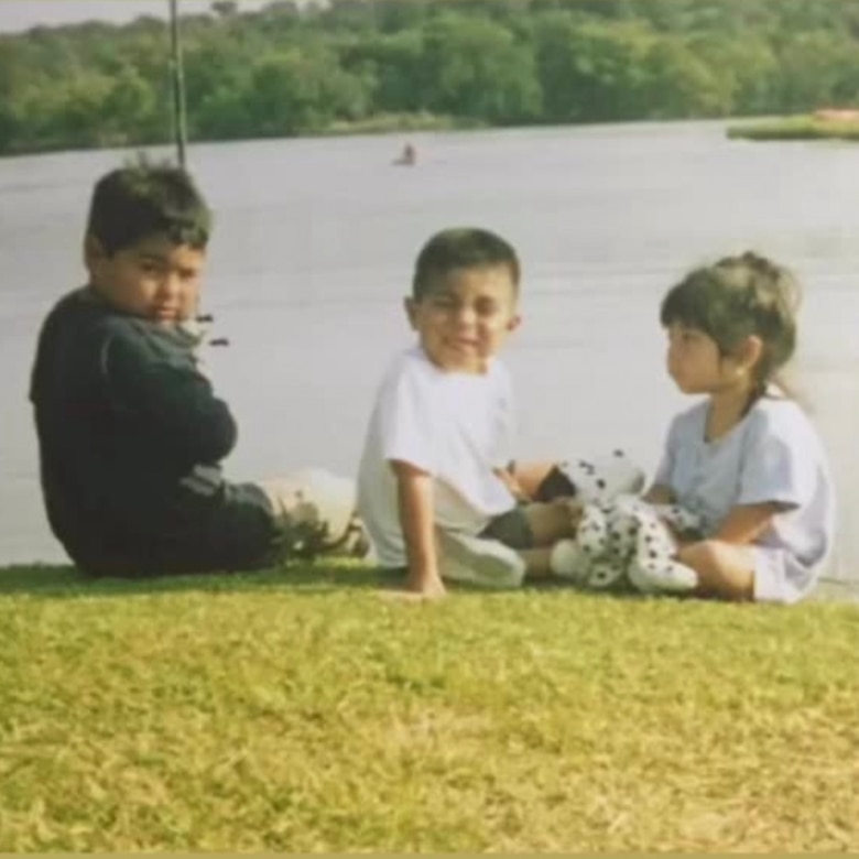 Three children sit on a grassy shoreline, one with a fishing pole.