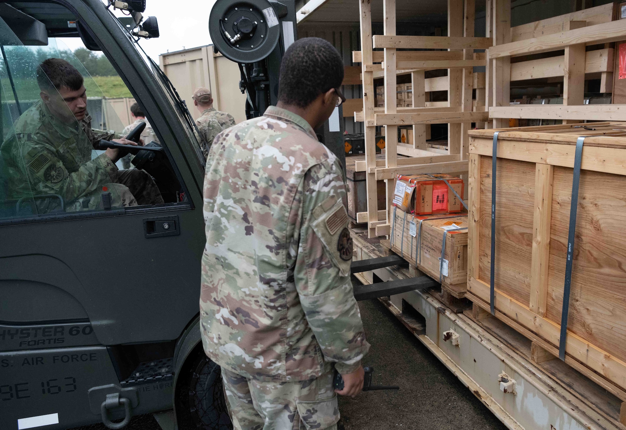 Two military members put a pallet in a shipping container. While one drives a forklift with the pallet the other guides him where to go.