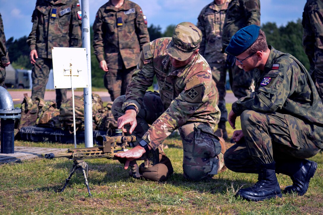 Two service members kneel on the ground to examine a weapon, as other soldiers stand in the background.