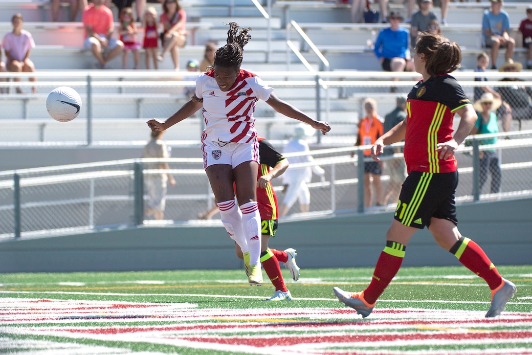 A soldier jumps to score a goal on a soccer field as another player observes.