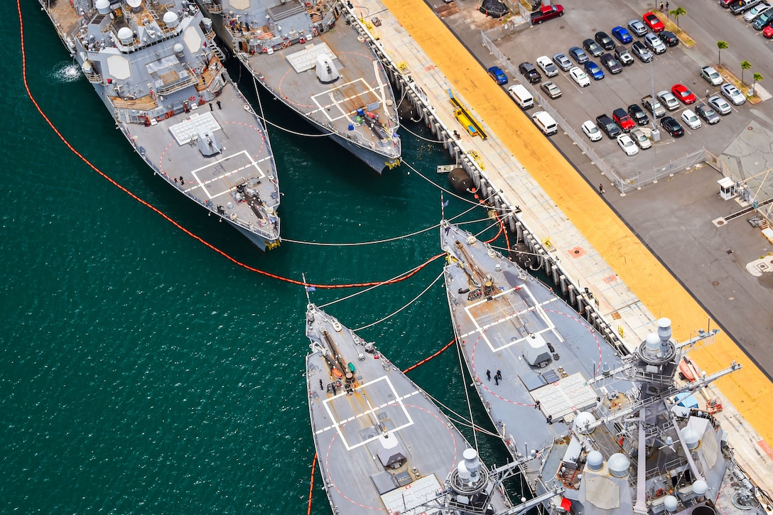 Four ships moored are seen from overhead alongside a pier with several parked vehicles.