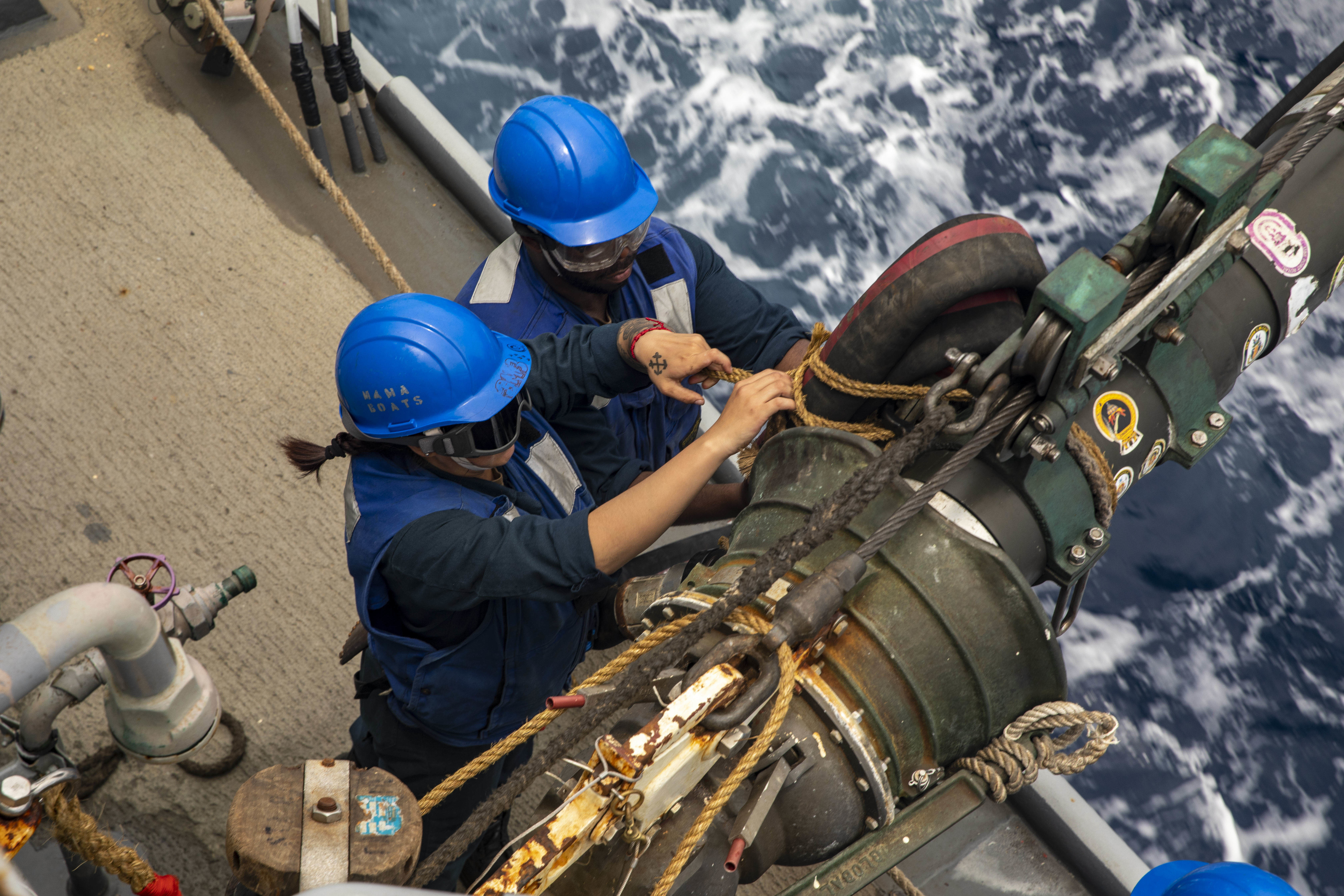 Two sailors connect a fuel line aboard a ship.