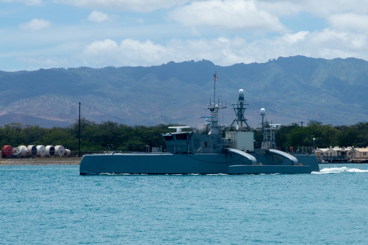 Waves break behind a vessel as it sails on the ocean; mountains are in the background.