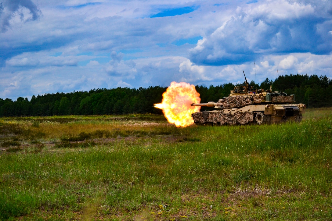 A military tank fires its weapon in an open grass field with trees in the background.