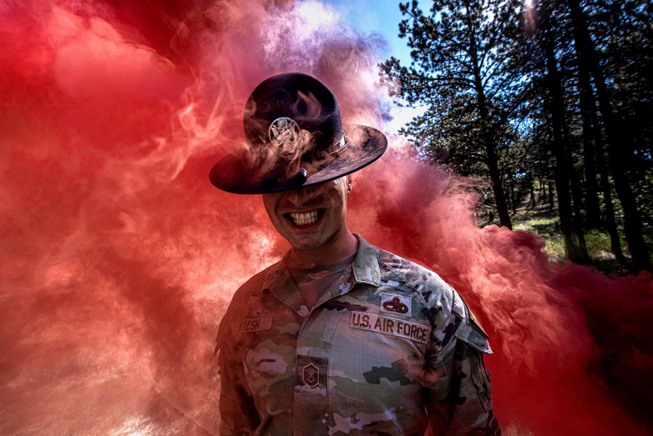 An airman smiles while surrounded by reddish smoke.