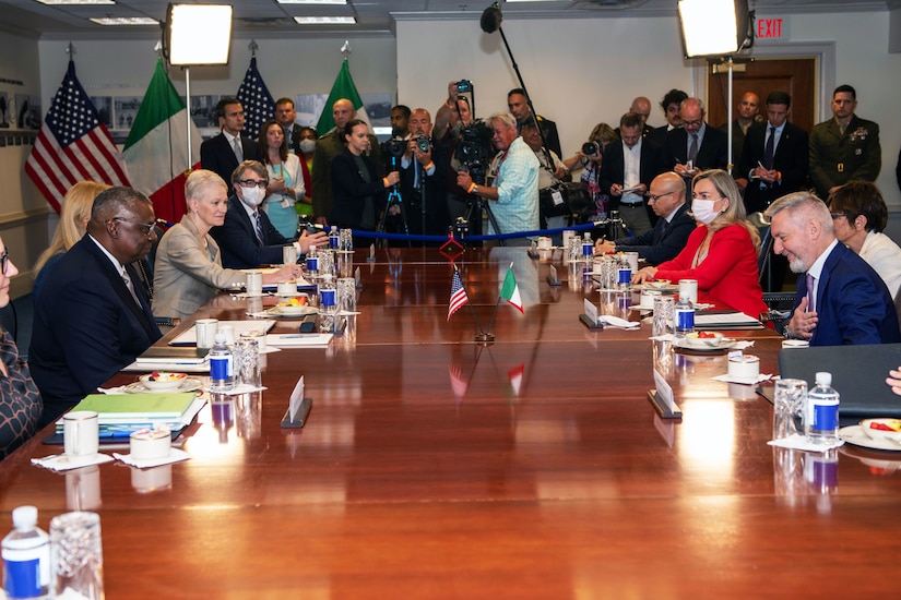 People in civilian attire are gathered around a large table. In the rear are members of the media. National flags are in the background.