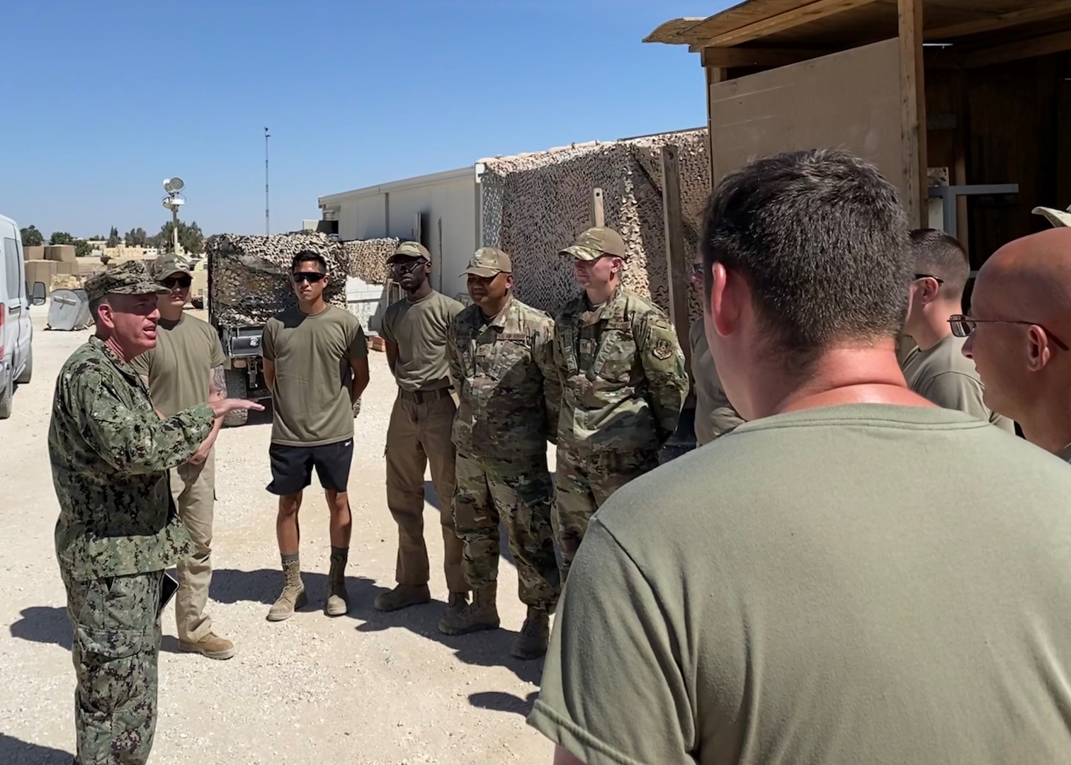 Man in camo uniform talks to a group of men in camo outdoors.