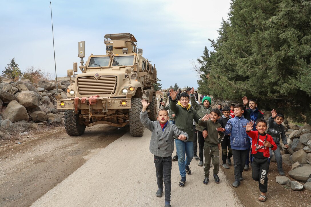 Children walk down a street. A large military vehicle is nearby.