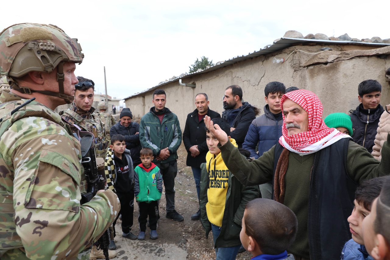 A uniformed service member talks with a civilian man. Children and other adults are nearby.
