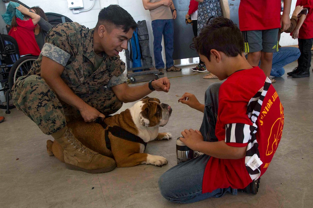 A Marine and a child kneel and hold treats above a resting bulldog.
