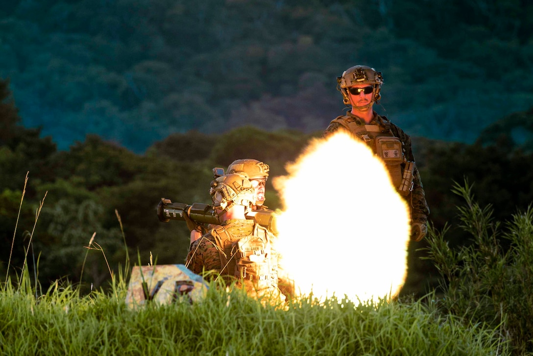 Marines fire a weapon as another Marine watches.