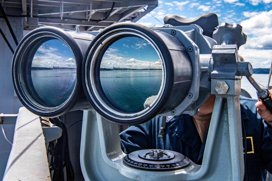 A sailor stands behind and looks through a large scope on a ship.