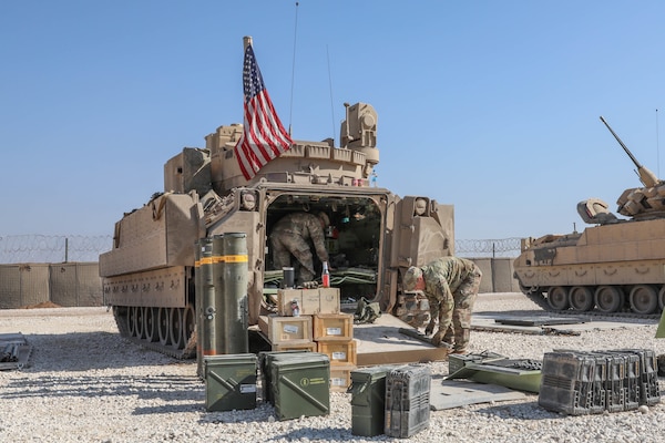 Two uniformed service members inside and outside a tank prepare it for combat. The tank displays an American flag.