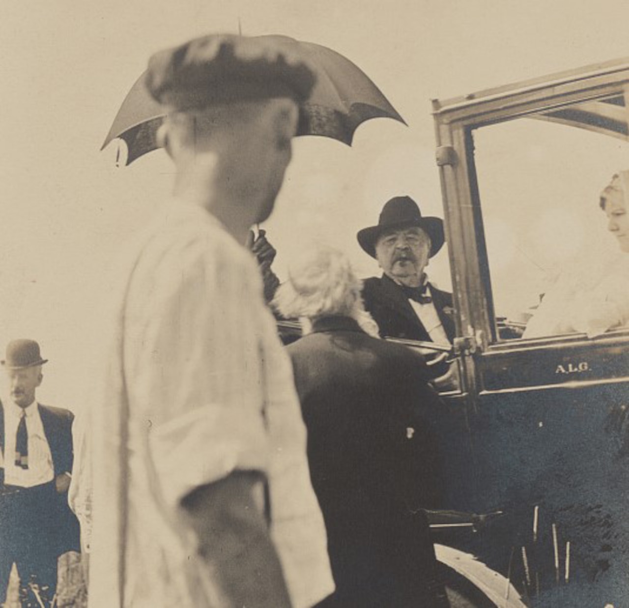 A man sits in an early-era vehicle while two others look at him.