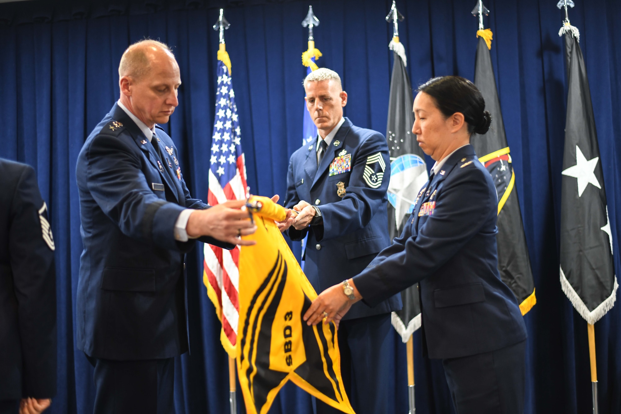 Col. Mia Walsh, commander, Space Base Delta 3, right, unfurls the SBD 3 guidon with Lt. Gen. Michael Guetlein, commander, Space Systems Command, left, on July 14 at Los Angeles Air Force Base.  Chief Master Sgt. Michael Groder, acting Senior Enlisted Leader for SBD 3, assists with the guidon.