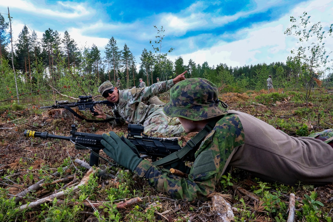 One soldier gestures to another soldier as the two aim their weapons in a clearing.