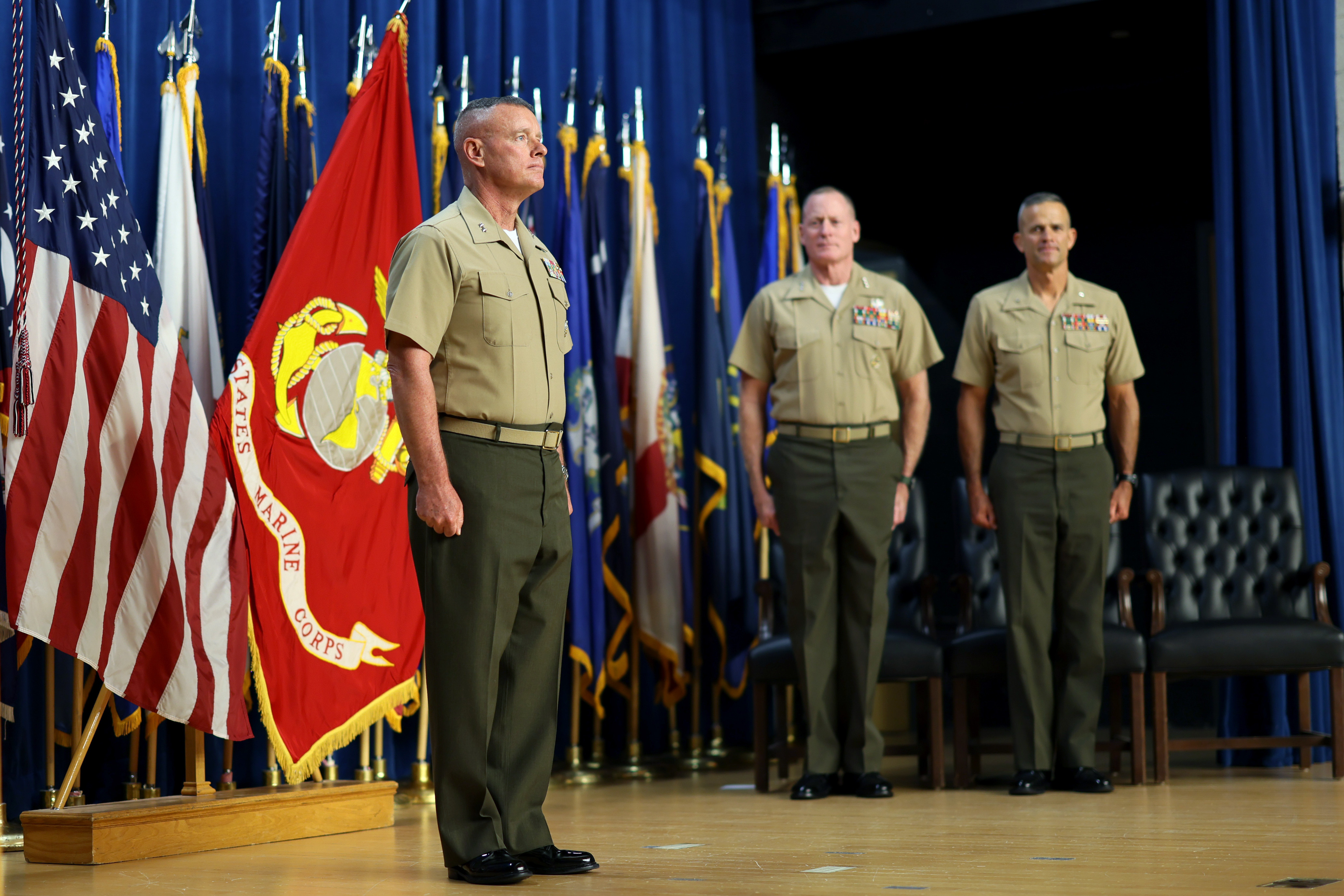 Four Marines on stage for a change of command ceremony. Various flags behind them.
