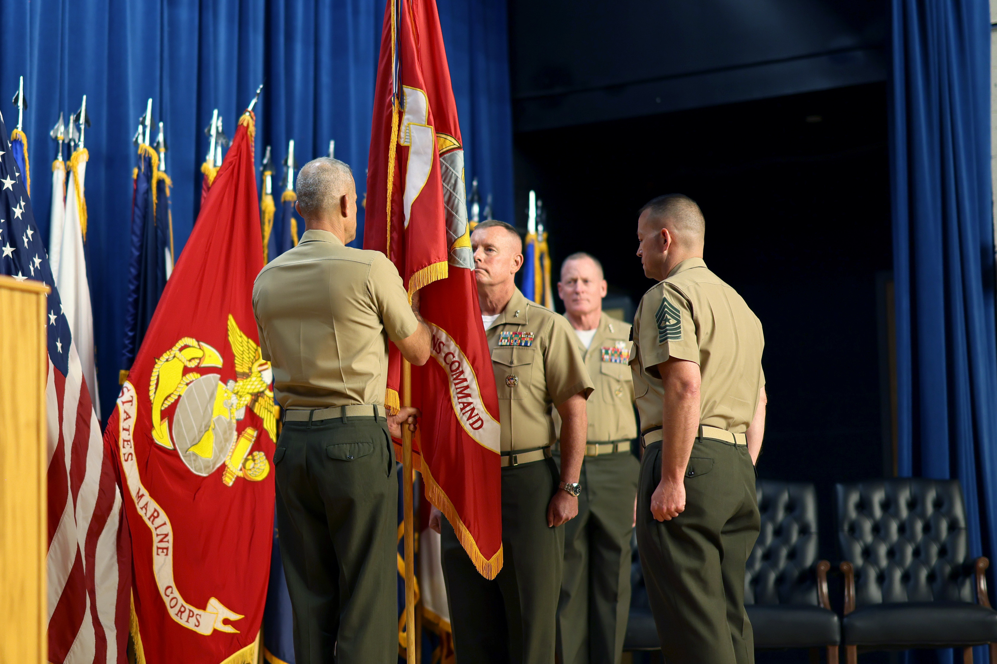 Four Marines on stage for a change of command ceremony. Various flags behind them.