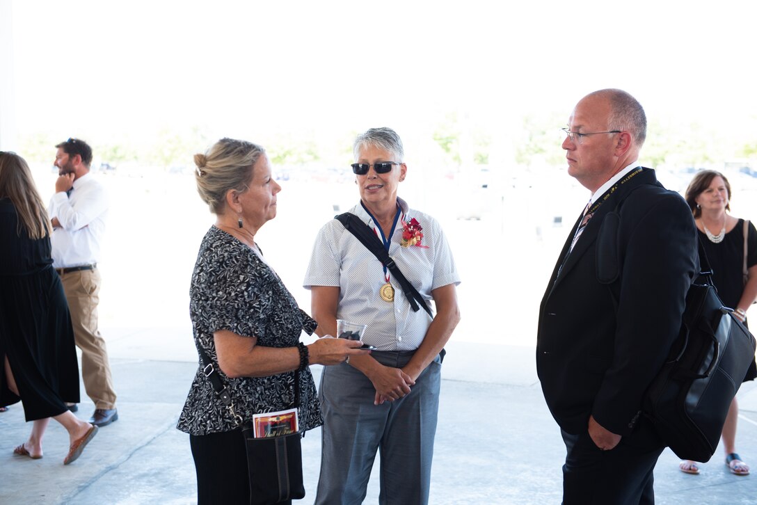A woman in a patterned black shirt and black pants with hair up in a bun chats with a woman in a white dress shirt and grey pants with a medal around her neck and a man in a dark dress suit under the shade of a pavilion outside.
