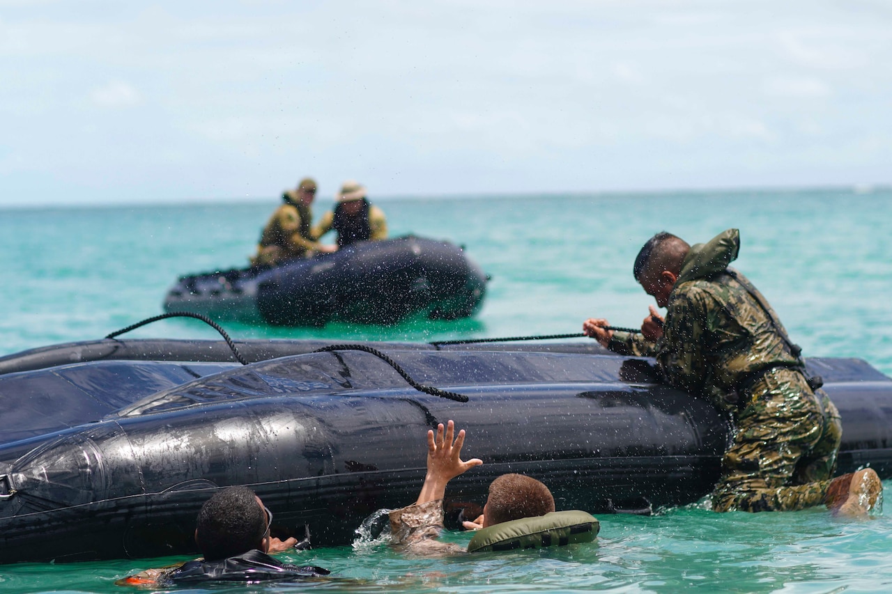 A Marine climbs on a flipped inflatable boat as two others swim beside the boat; another inflatable boat with two people on board can be seen in the background.