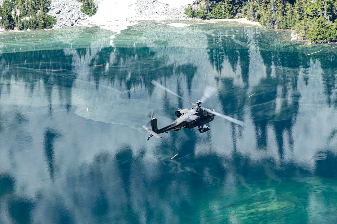 An Army helicopter flies over bluish waters reflecting trees and mountains.