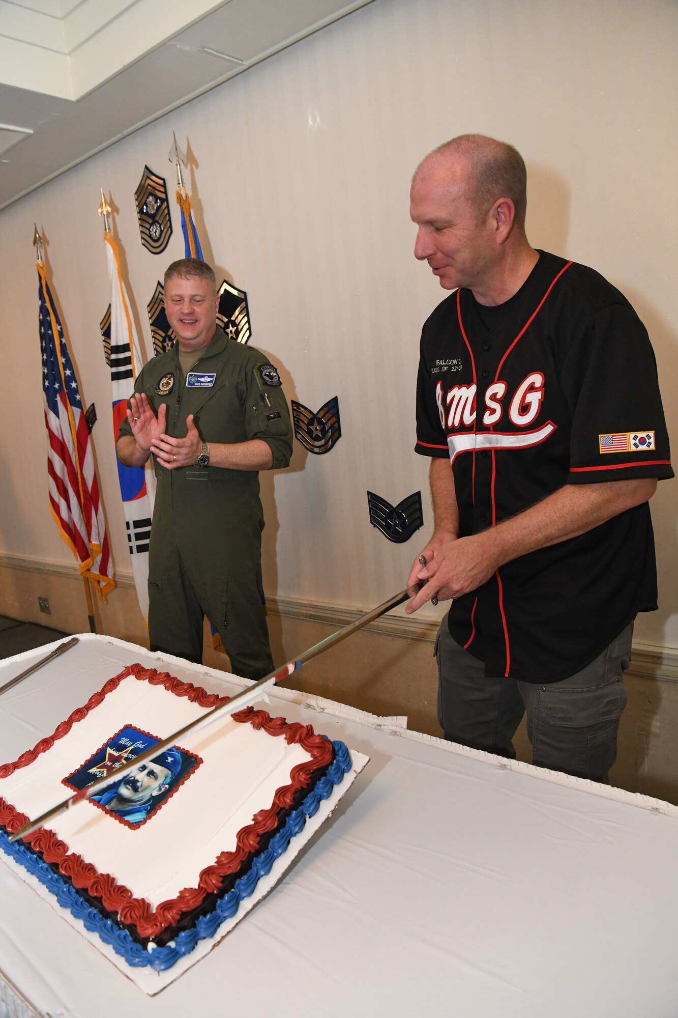 A military member watches while a military member wearing a jersey cuts a cake with a sword.