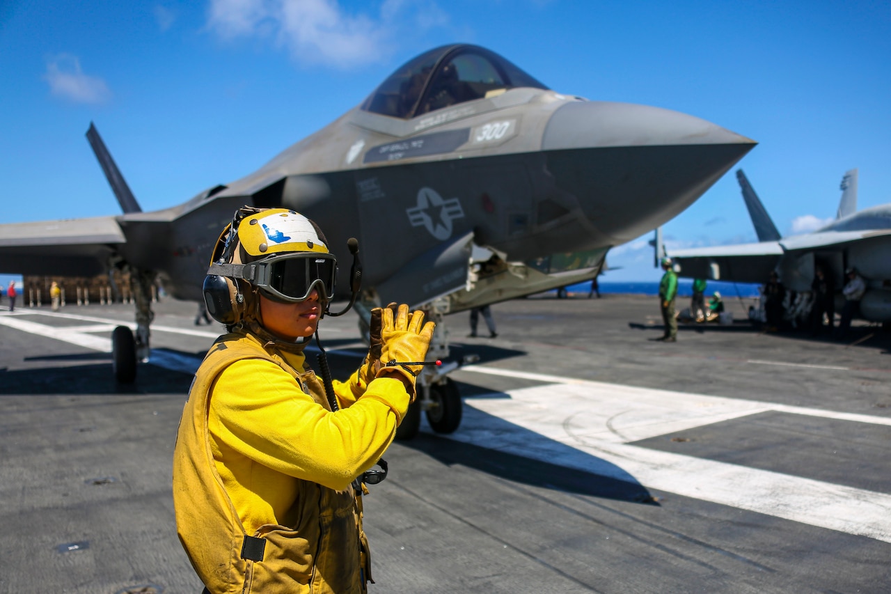 A sailor in bright yellow gives hand signals to the pilot of an aircraft in a deck.