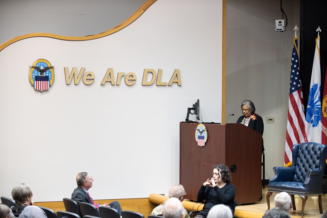 A woman in black speaks at a podium on stage.