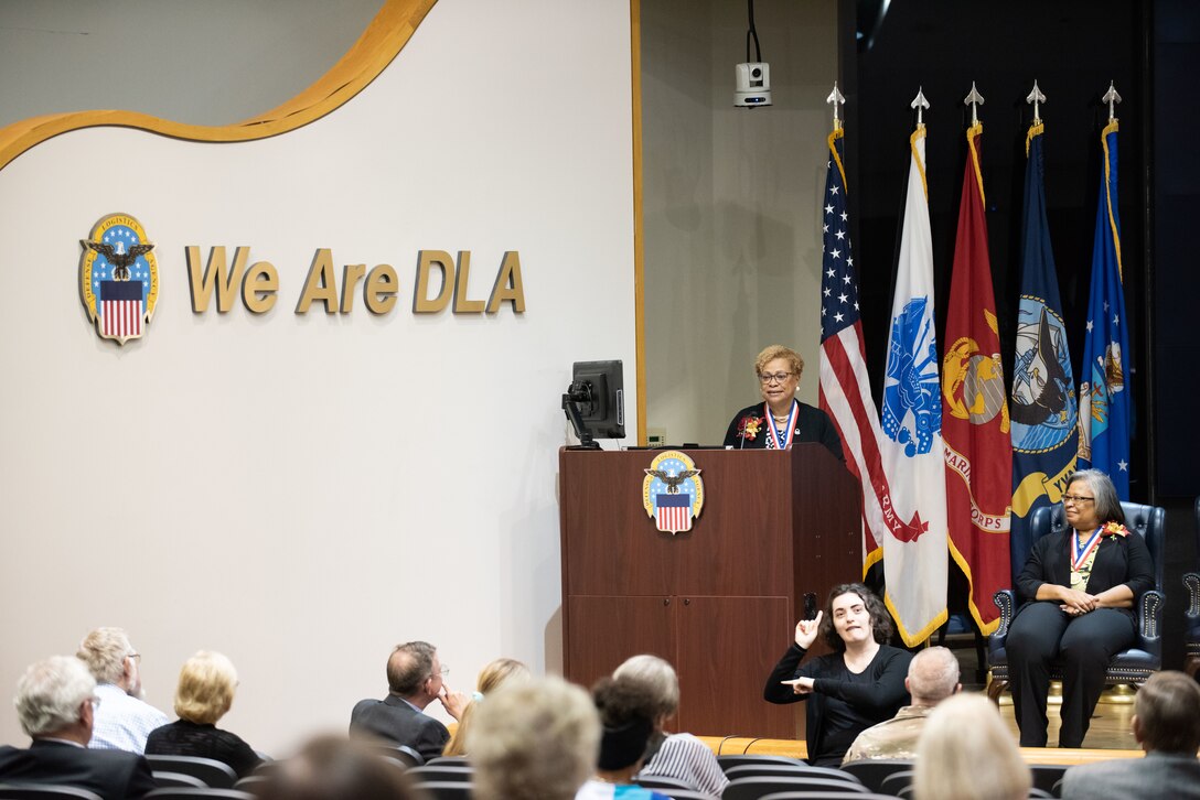 A woman in a black patterned dress speaks at a podium on stage.