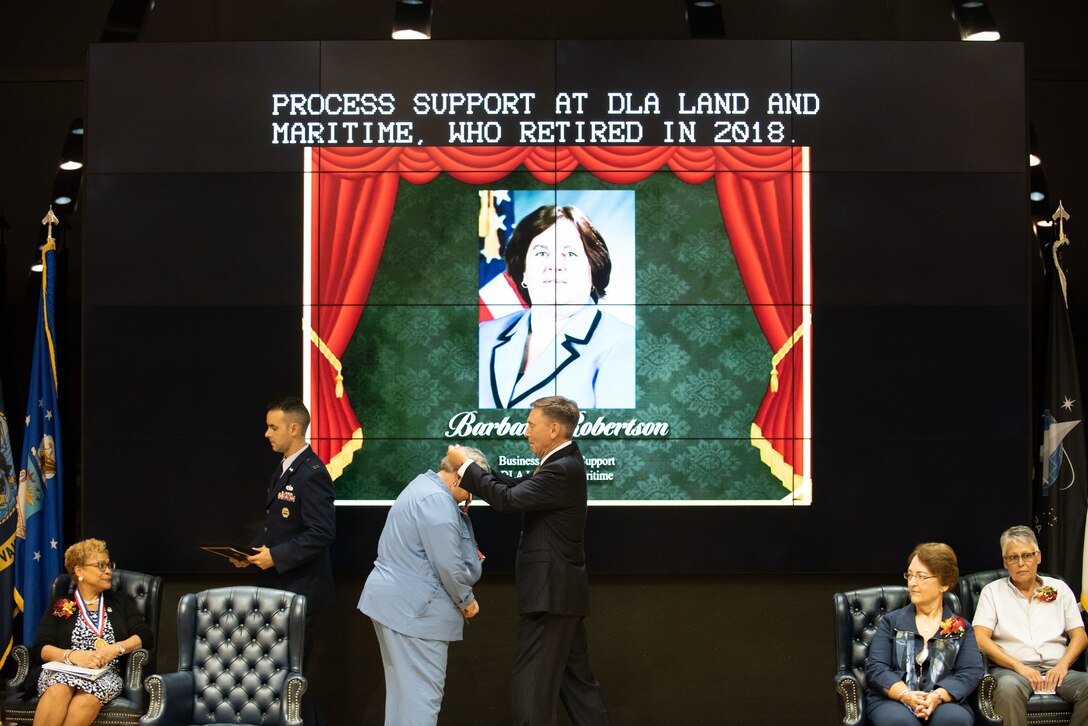 A woman in a blue suit gets a medal placed around her neck by a man in a dark suit.