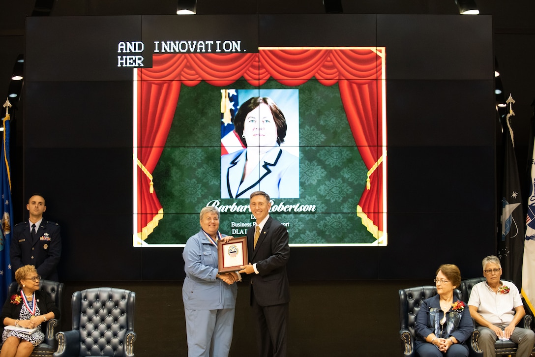 A woman in blue holds a plaque with a man in a dark suit on stage.