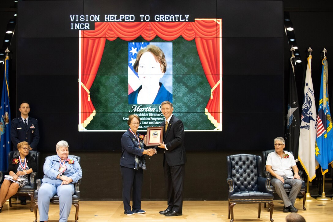 A woman dressed in blue holds a plaque on stage with a man in a dark suit.