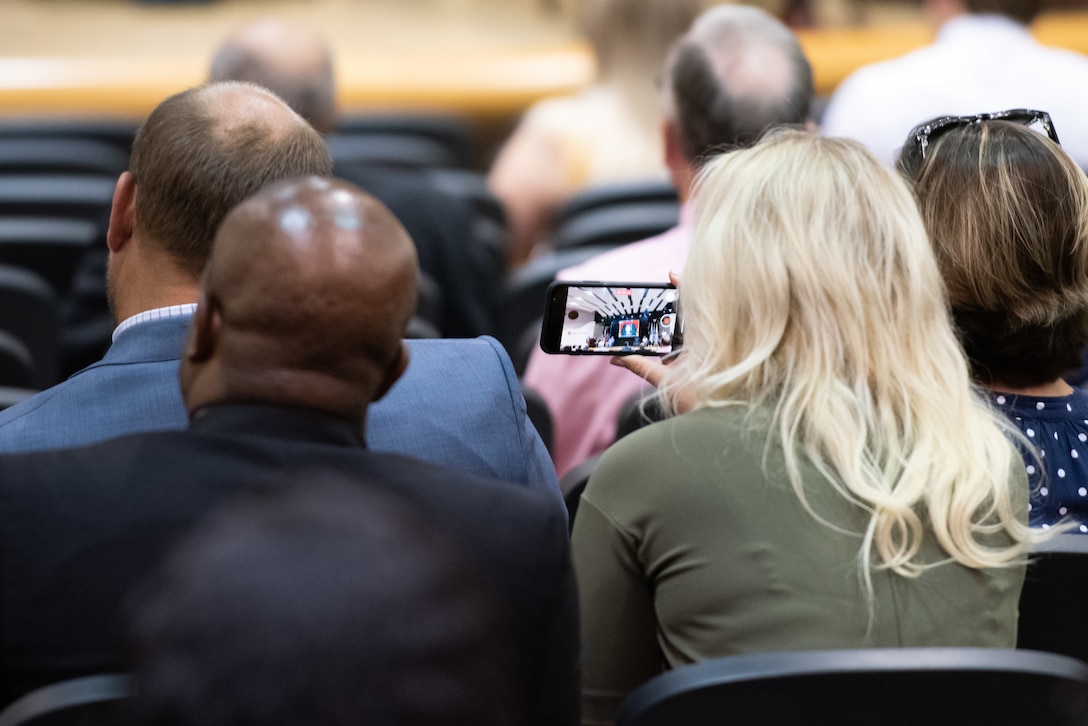 A man and a woman seated in a auditorium. The woman shoots video on her phone.
