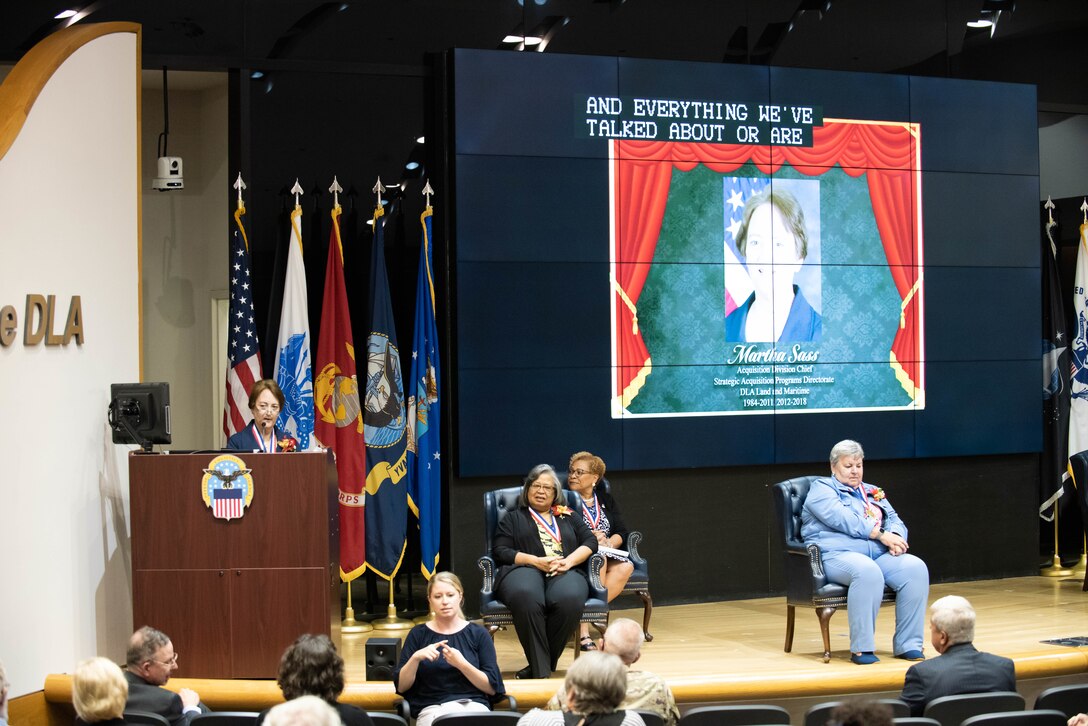 A woman in blue speaks at a podium on stage with three other women sitting on stage listening.