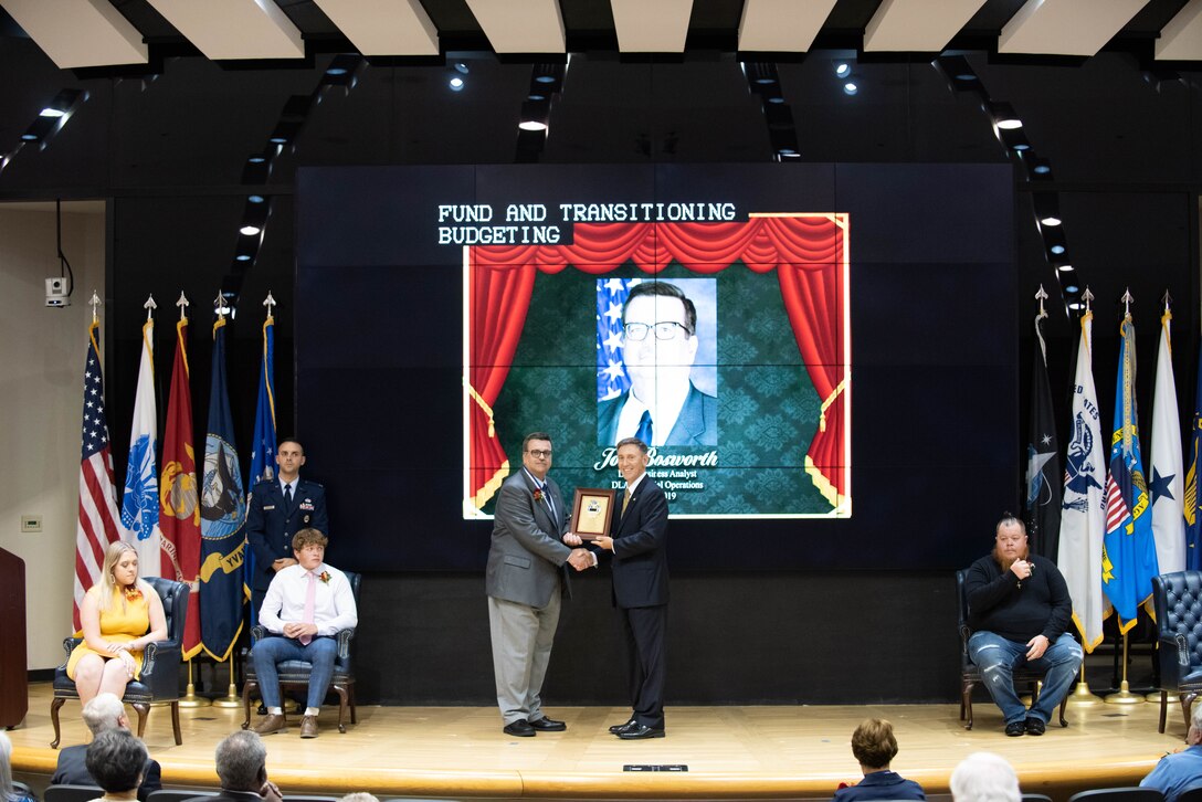 A man in a light grey suit holds a plaque with a man in a dark grey suit on stage.