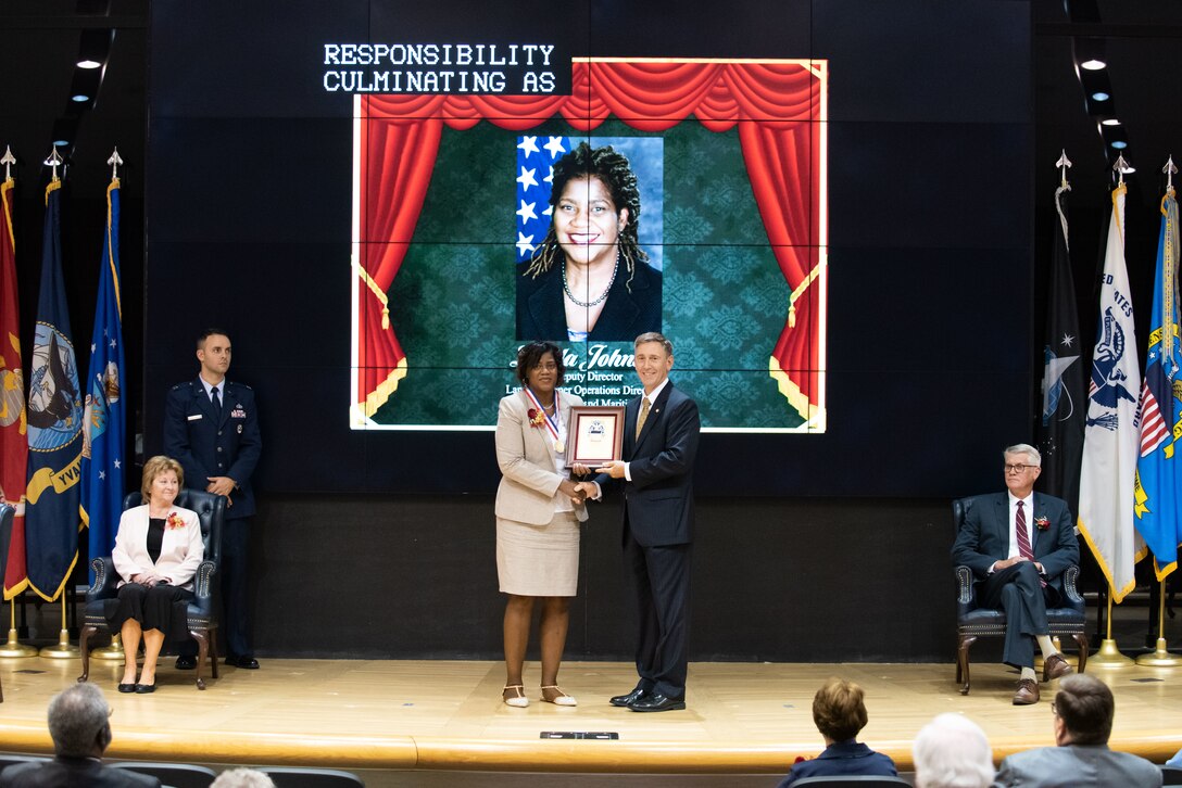 A woman in a tan suit and a man in a grey suit hold a plaque on stage.