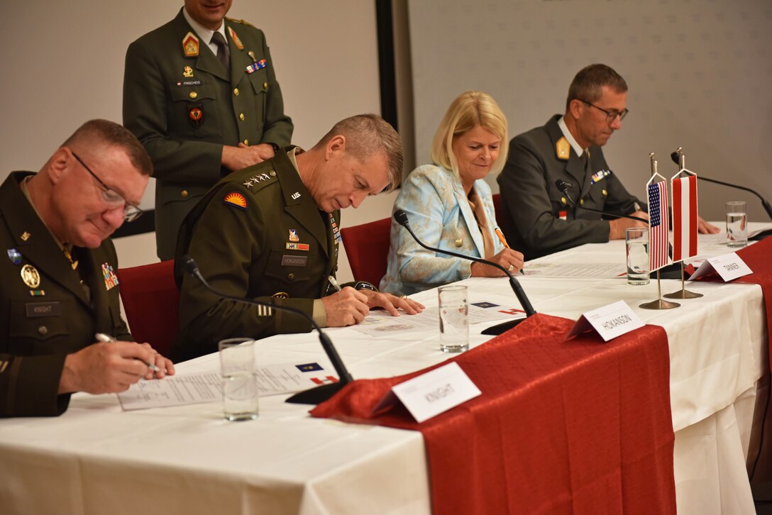 A military person supervises as four people seated at a table sign documents.