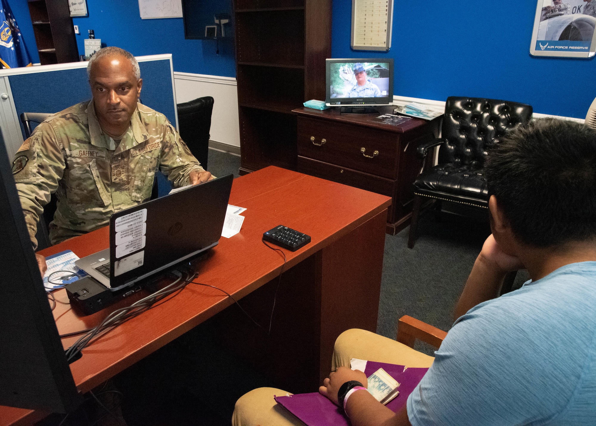 MSgt. Gaffney sits behind his desk looking at a computer while a trainee sits on the other side of the desk watching a tv in the background