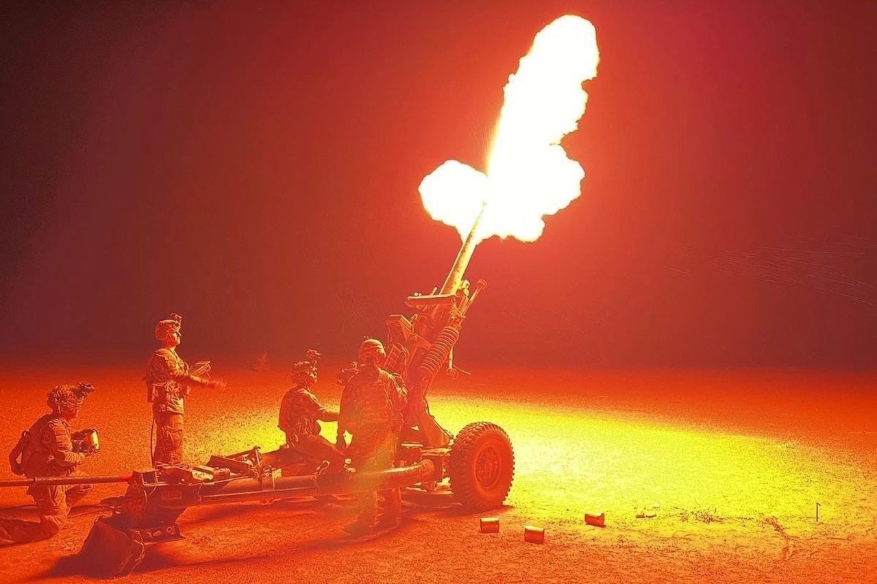 Four service members observe large artillery as its fire power light’s up the night sky.