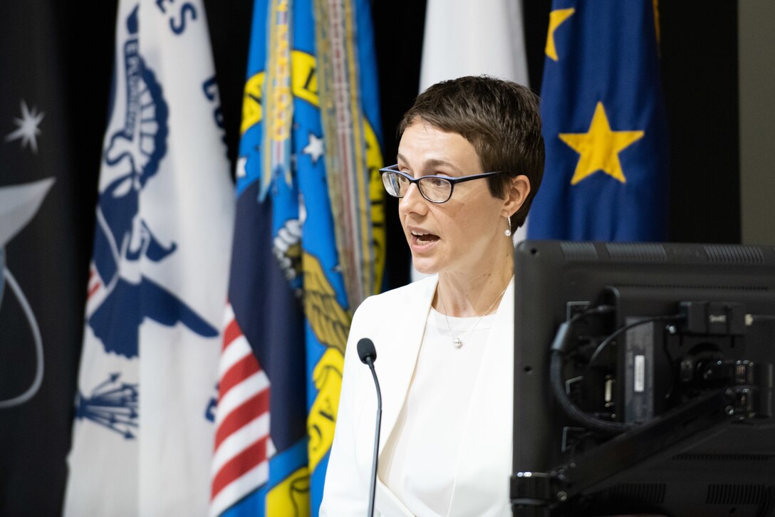 A woman in a white suit and short hair speaks at a podium in an auditorium.