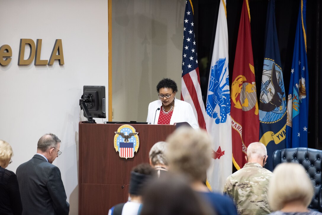 A woman in a white suit speaks at a podium in an auditorium.