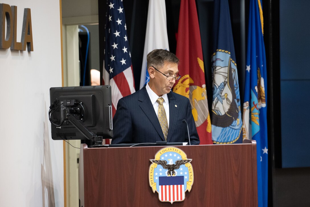 A man in a grey suit at a podium on a stage in a auditorium.