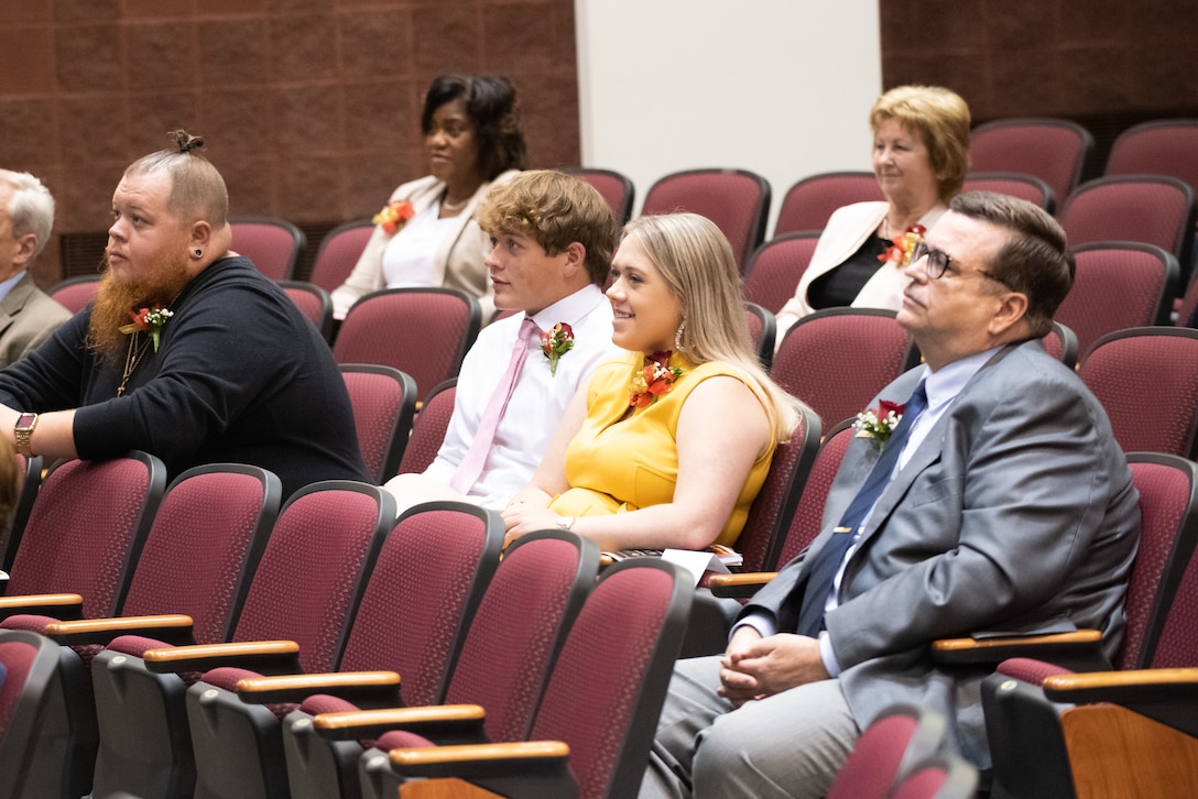 A man in a black shirt, a man in a white dress shirt, a woman in a yellow dress and a older man in a grey suit sit in a auditorium.